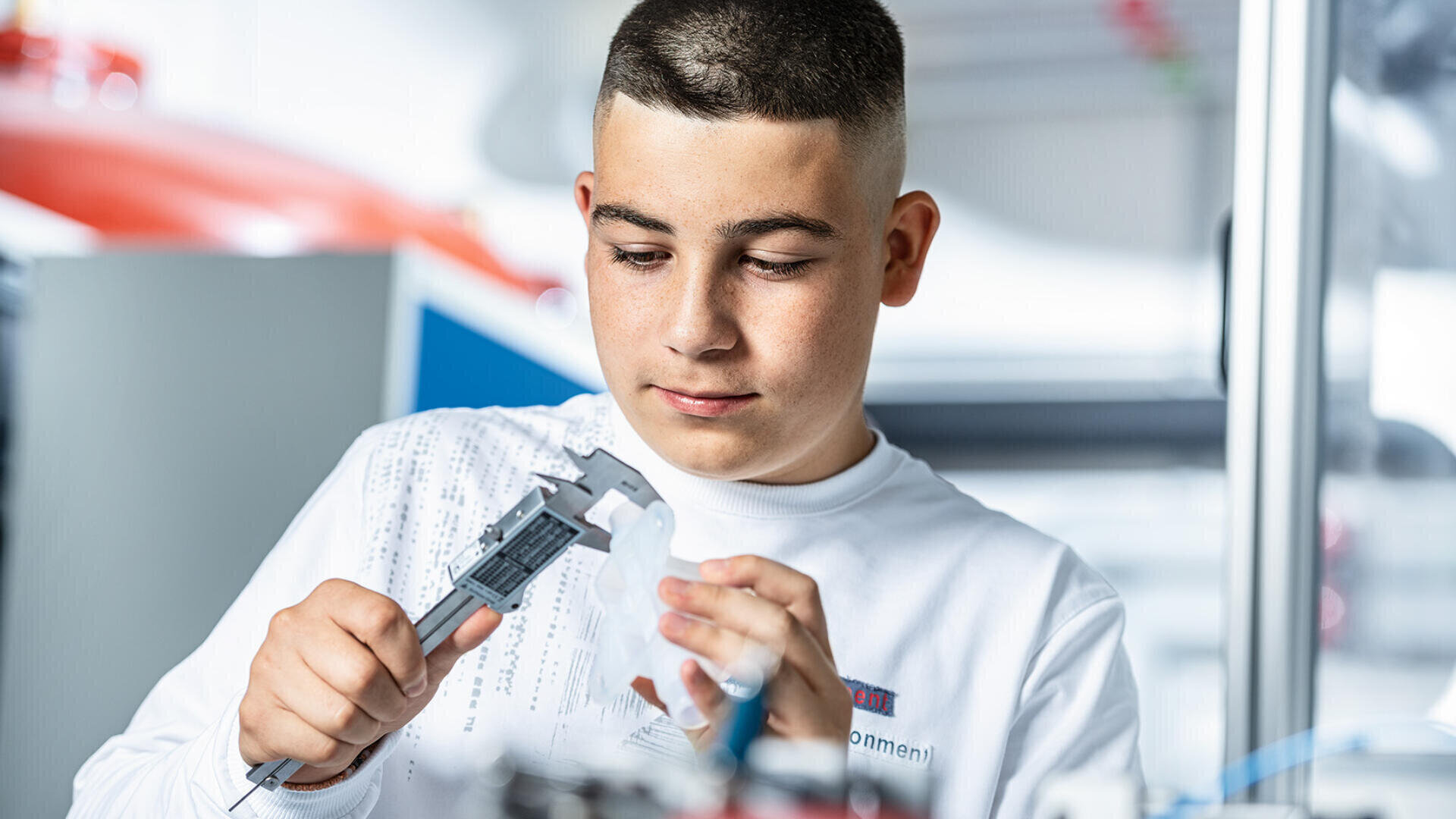 Ein junger Mensch in einem weißen Hemd misst mit einem digitalen Messschieber einen kleinen Gegenstand in einem hell erleuchteten, modernen Arbeitsraum.