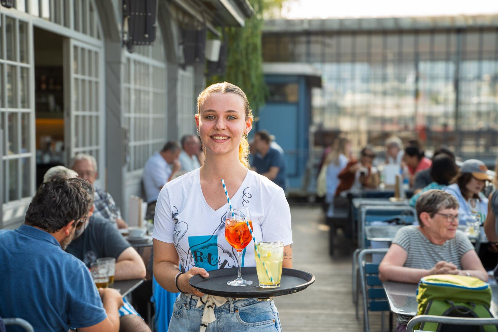 Eine lächelnde Kellnerin hält ein Tablett mit zwei farbenfrohen Getränken auf einer Terrasse im Freien, umgeben von Menschen, die an Tischen sitzen und Essen und Getränke im Sonnenlicht genießen.