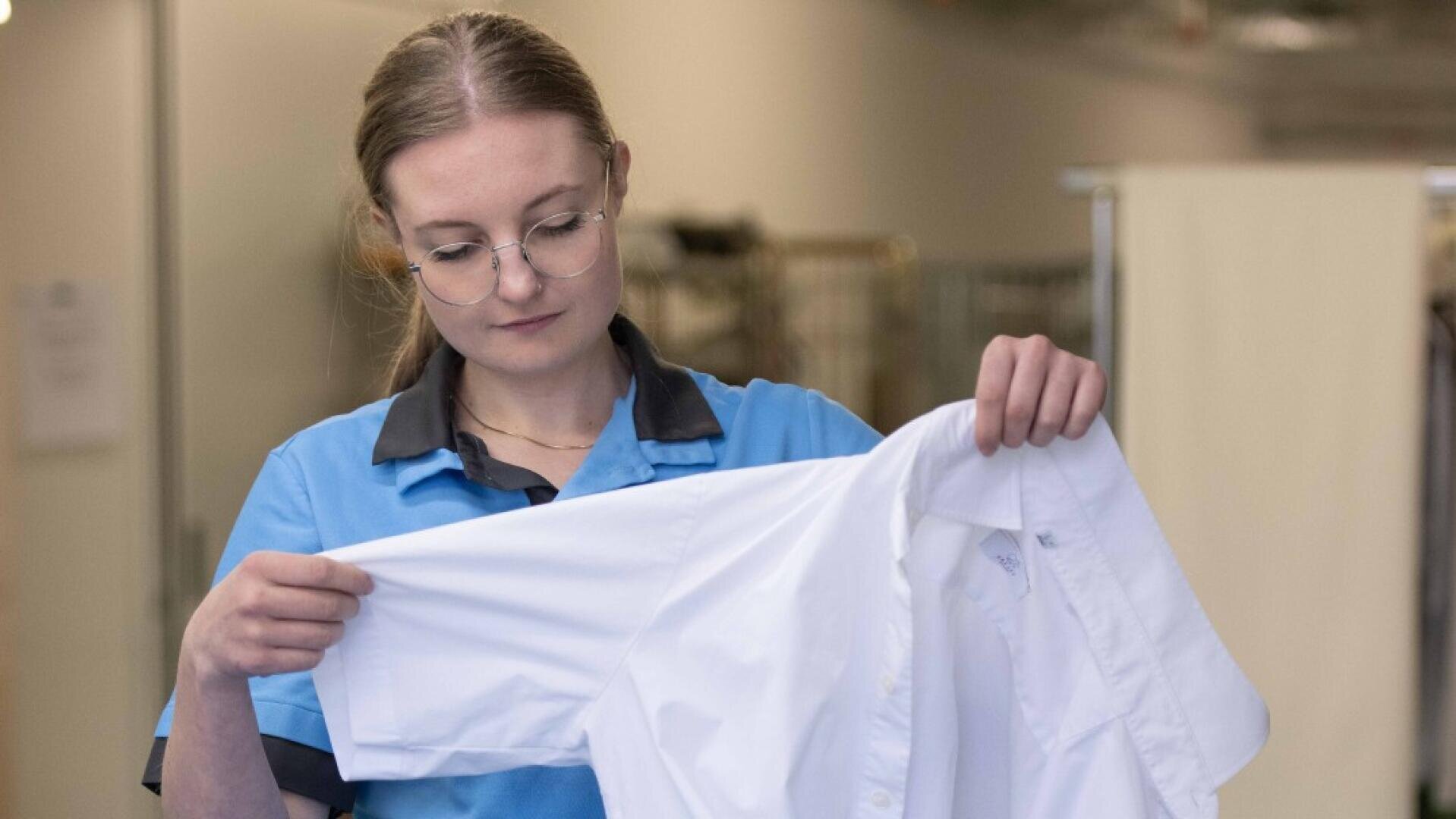 Eine Frau mit Brille und blauer Uniform inspiziert in einem gut beleuchteten Innenraum ein weißes Hemd, indem sie es hochhält und den Ärmel genau betrachtet.