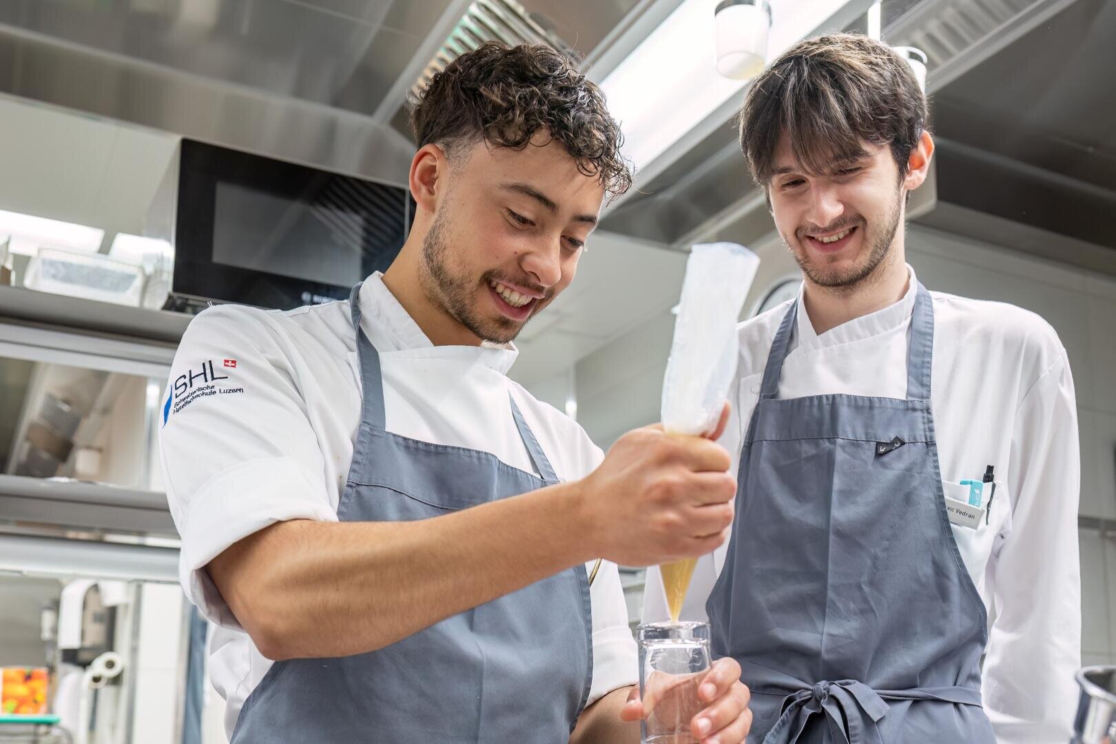Zwei junge Köche in weißen Uniformen und grauen Schürzen lächeln, während der eine in einer professionellen Küche eine Mischung in ein Glas füllt.