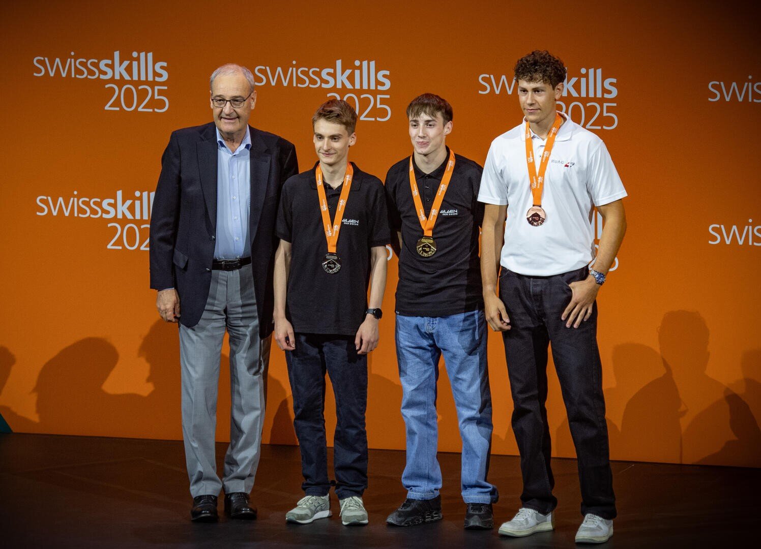 Vier MĂ€nner stehen vor einer orangefarbenen Kulisse der SwissSkills 2025; drei junge MĂ€nner mit Medaillen und Freizeitkleidung stehen neben einem Ă€lteren Mann im Anzug, alle lĂ€cheln fĂŒr ein Gruppenfoto.