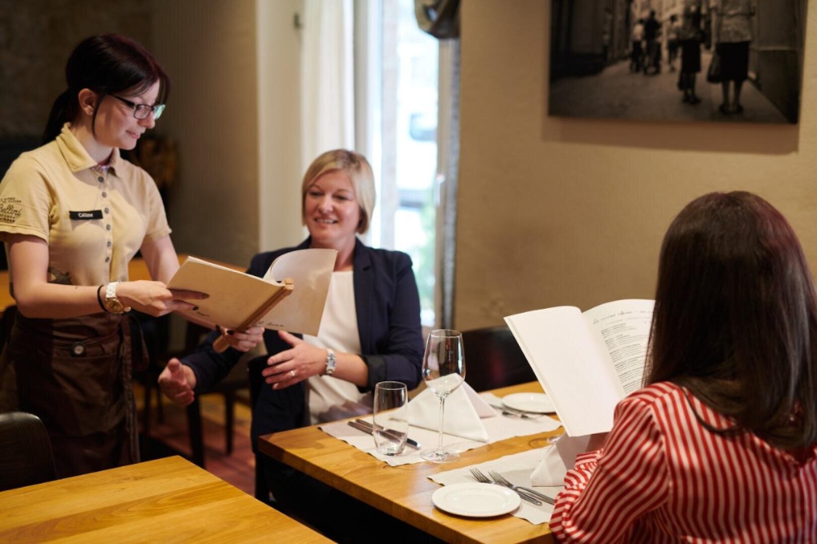 Eine Kellnerin reicht einer lächelnden Frau, die an einem Tisch im Restaurant sitzt, eine Speisekarte, während eine andere Frau in einem rot gestreiften Hemd ihre Speisekarte ansieht.