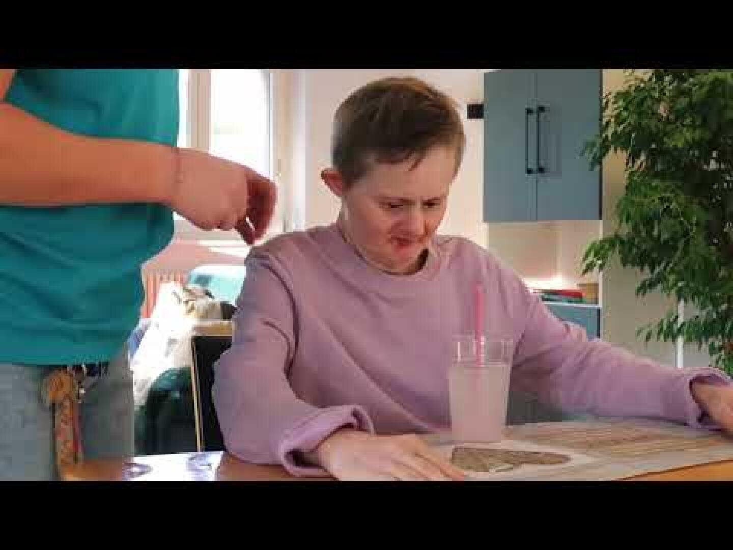 Eine Person mit Down-Syndrom sitzt an einem Tisch mit einem Getränk und einem Strohhalm und macht einen unzufriedenen oder unangenehmen Gesichtsausdruck. Eine andere Person steht daneben, teilweise sichtbar.