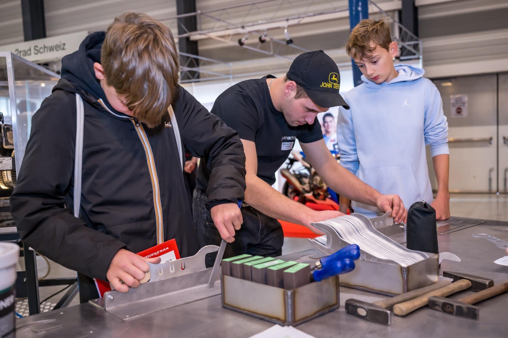 Drei junge Menschen stehen an einer Werkbank und hantieren mit Metallwerkzeugen und -teilen. Einer konzentriert sich auf ein Metallteil, ein anderer schaut nach unten, und der dritte beobachtet. Der Schauplatz scheint ein Industrie- oder Werkstattbereich zu sein.