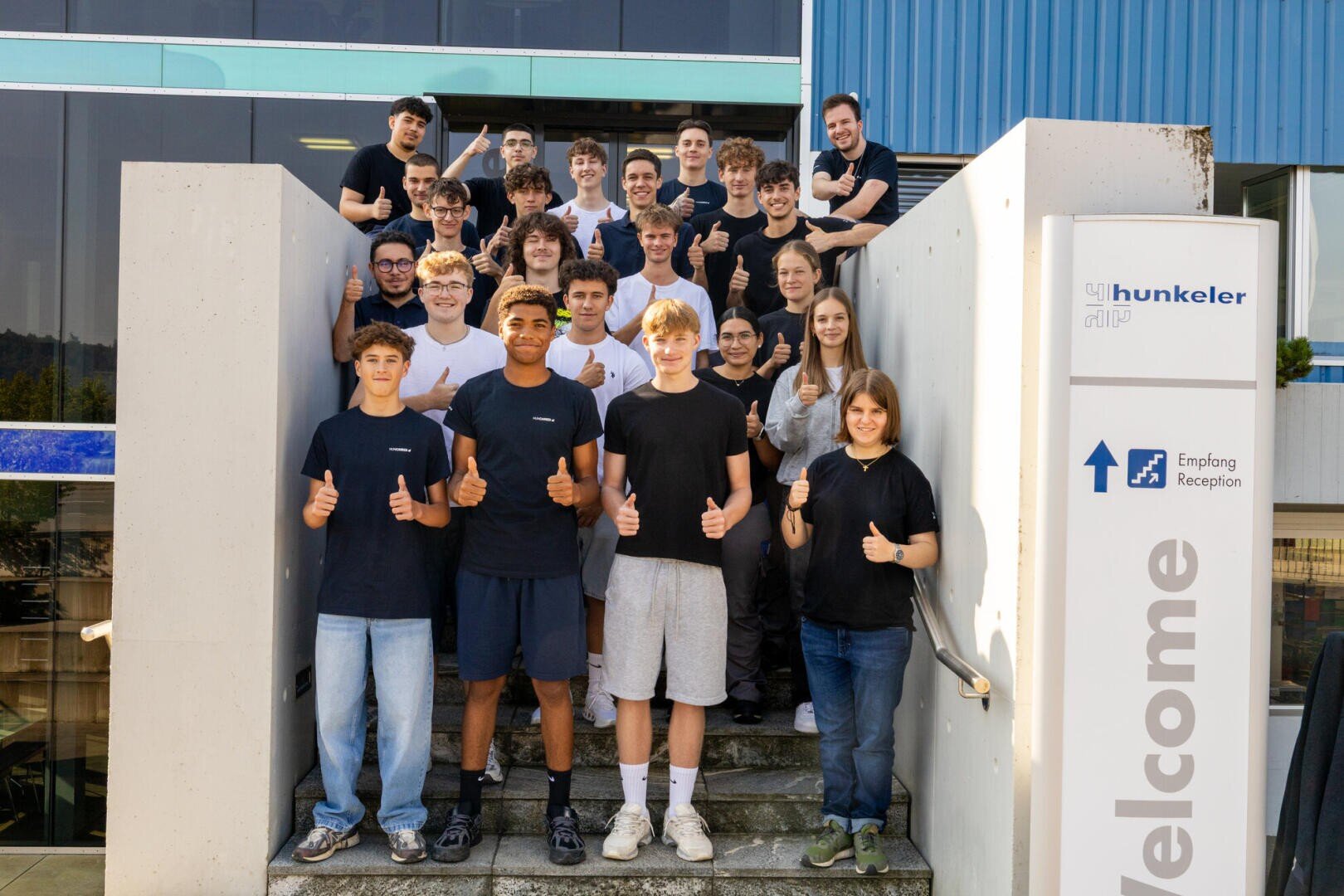 Eine Gruppe junger Menschen, meist Teenager, steht auf einer Freitreppe vor einem modernen Gebäude, lächelt und zeigt der Kamera die Daumen nach oben. Im Vordergrund ist ein großes Willkommensschild zu sehen.