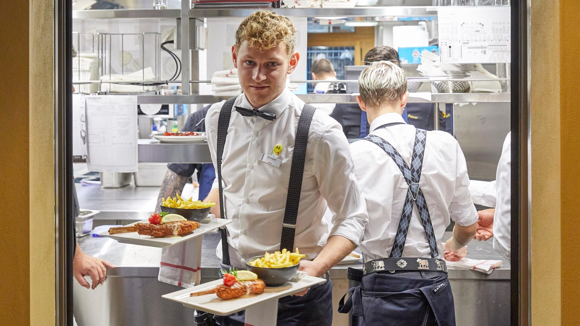 Ein Kellner mit weißem Hemd, Fliege und Hosenträgern trägt Teller mit Essen und Pommes frites, während das Küchenpersonal im Hintergrund in einer geschäftigen Restaurantküche arbeitet.