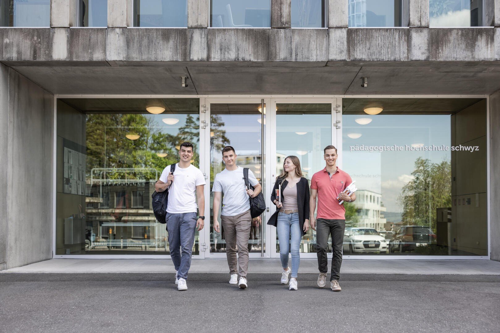 Vier junge Erwachsene, drei Männer und eine Frau, treten mit Taschen und Büchern aus einem modernen Gebäude mit großen Glastüren. Auf dem Fenster des Gebäudes steht "pädagogische hochschule schwyz".