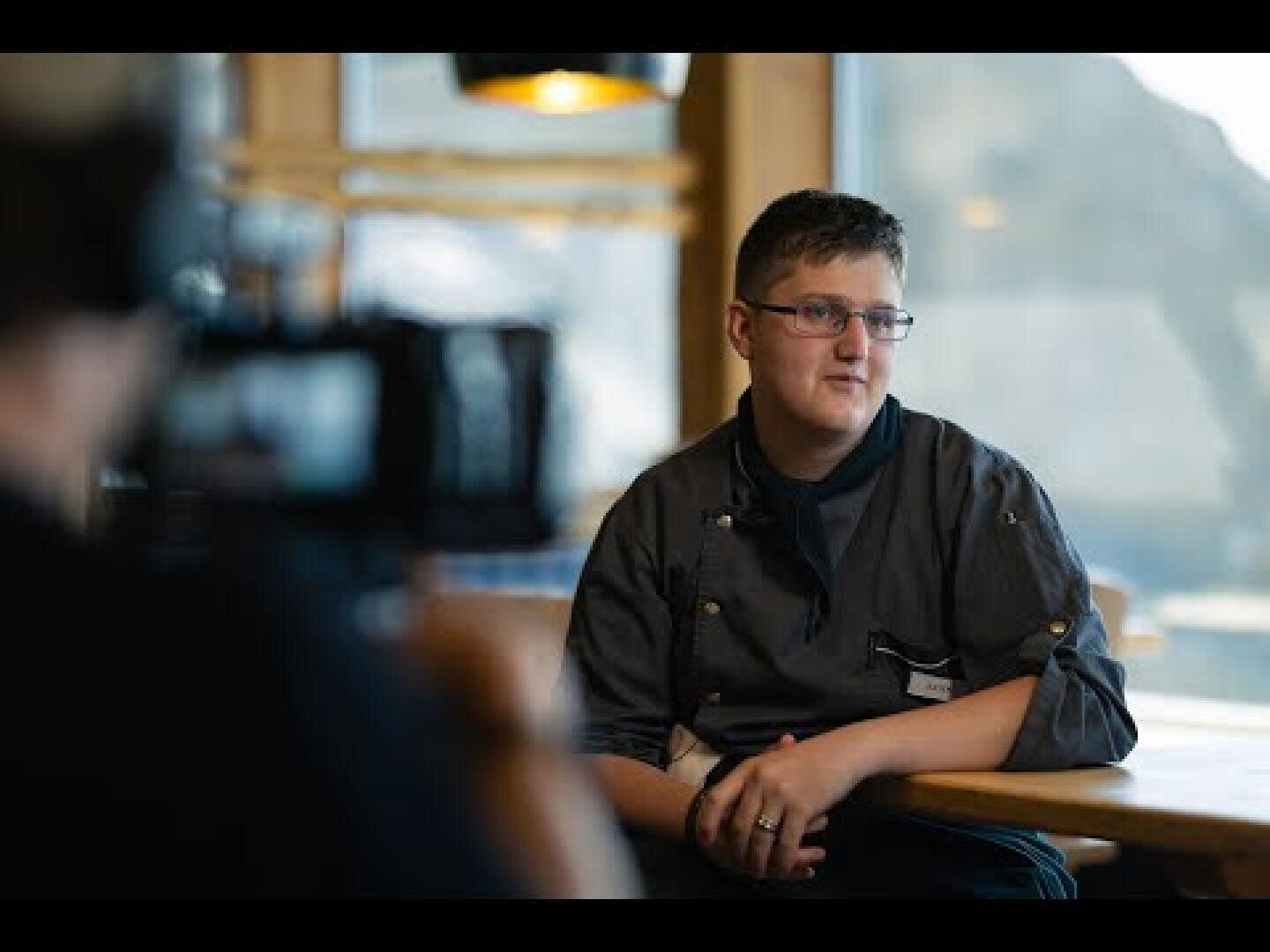 Eine Person mit Brille und Kochjacke sitzt an einem Holztisch in einem gut beleuchteten Raum und blickt leicht zur Seite. Eine verschwommene Kamera und eine weitere Person sind im Vordergrund zu sehen.
