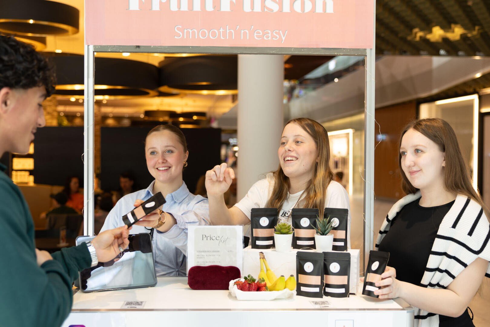 Drei junge Frauen stehen hinter einem Stand mit der Aufschrift FruitFusion, lächeln und reichen einem männlichen Kunden Getränke. Becher und frisches Obst sind auf dem Tresen in einem hellen Innenraum angeordnet.