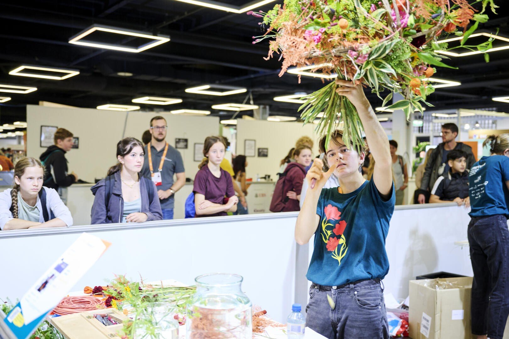 Eine Person hält einen großen Blumenstrauß in die Höhe, während sie einer Gruppe von aufmerksamen Zuschauern in einem hell erleuchteten Innenraum ein Blumenarrangement vorführt.