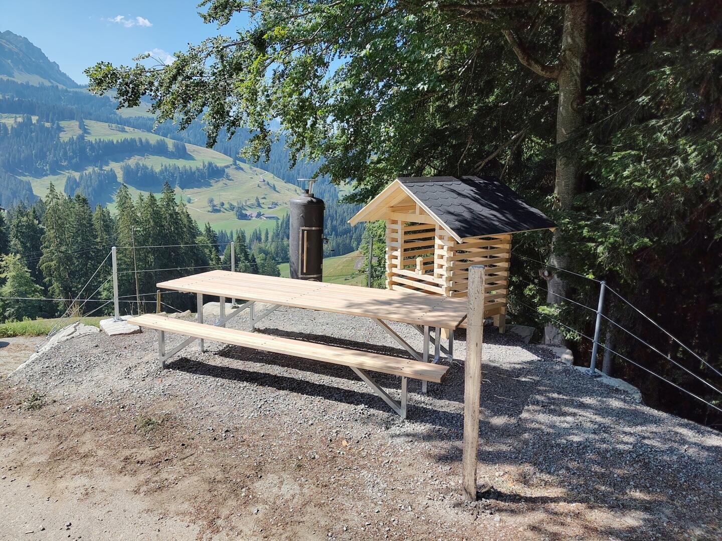 Auf einem Kiesplatz stehen ein Picknicktisch aus Holz und ein kleiner Unterstand aus Holz, umgeben von Bäumen und Bergen unter blauem Himmel. In der Nähe steht ein Metallcontainer, im Hintergrund sind malerische Hügel zu sehen.