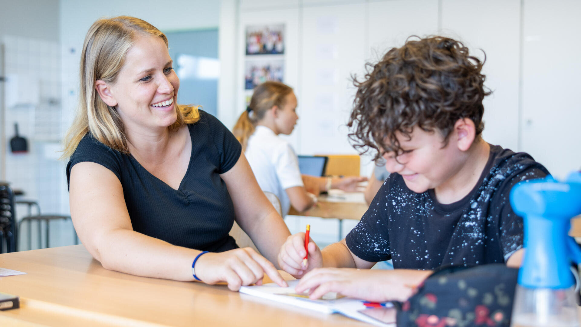 Ein lächelnder Lehrer sitzt neben einem Schüler, der in ein Notizbuch schreibt. Die Lehrerin zeigt auf das Papier und gibt Hilfestellung, während ein anderer Schüler im Hintergrund arbeitet. Der Schauplatz ist ein helles Klassenzimmer.