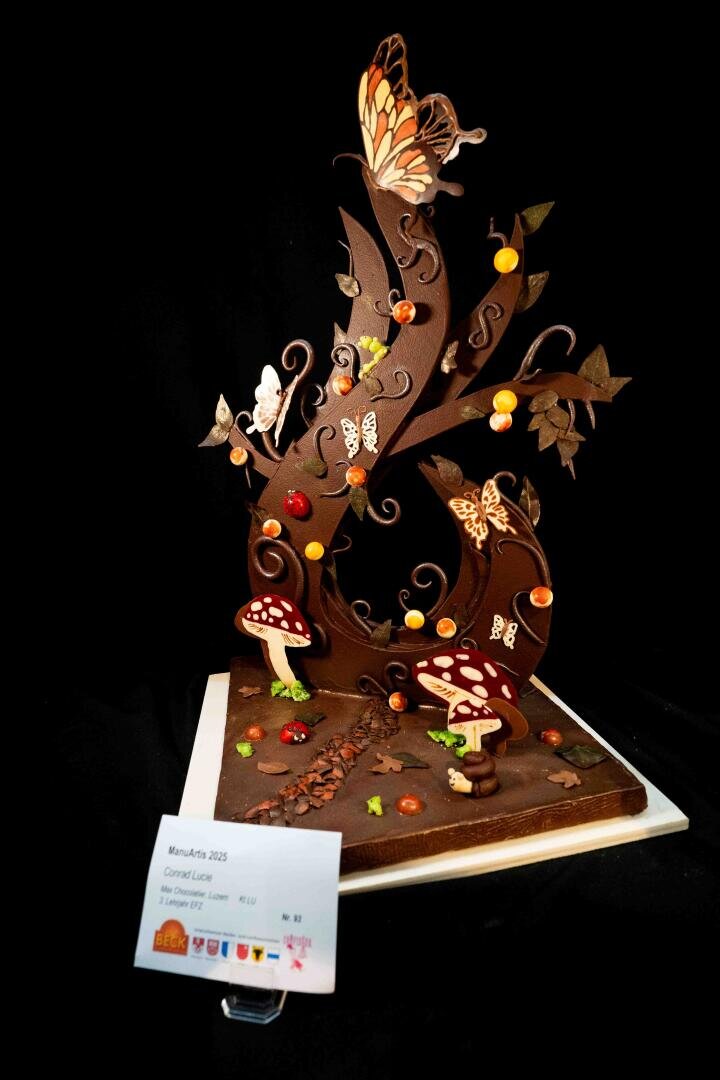 Eine Schokoladenskulptur mit verspielten Schnörkeln, Schmetterlingen, Pilzen und bunten Bonbons steht auf einem quadratischen Sockel, vor dem eine Beschriftungstafel angebracht ist. Der schwarze Hintergrund hebt die Details der Skulptur besonders hervor.
