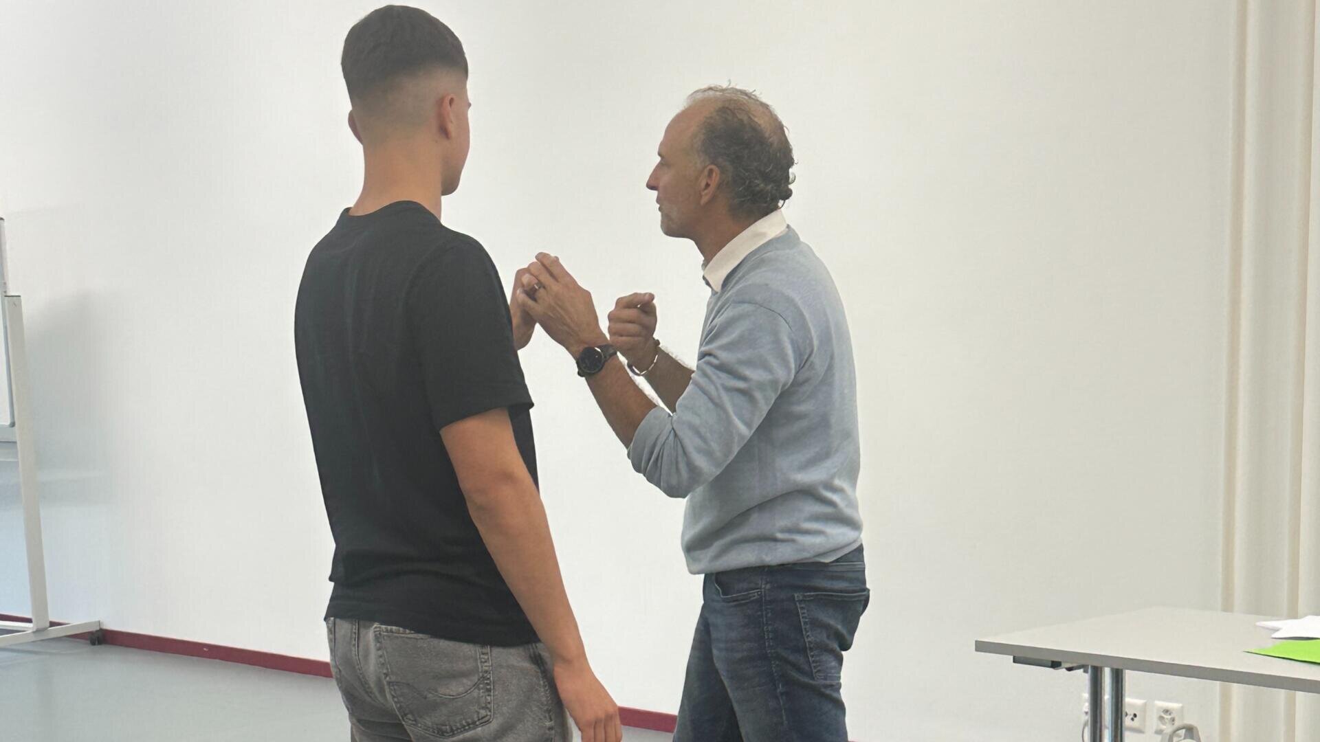Zwei Männer stehen sich in einem Innenraum gegenüber; der eine trägt ein schwarzes Hemd und Jeans, der andere einen hellblauen Pullover und Jeans, und es sieht so aus, als würden sie gemeinsam eine Körpertechnik demonstrieren oder üben.
