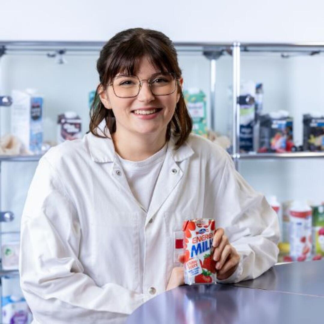 Eine Frau in einem weißen Laborkittel und mit Brille lächelt, während sie in einem Labor einen Karton Energy Milk mit Erdbeergeschmack hält, während auf Regalen hinter ihr verschiedene Getränkekartons ausgestellt sind.