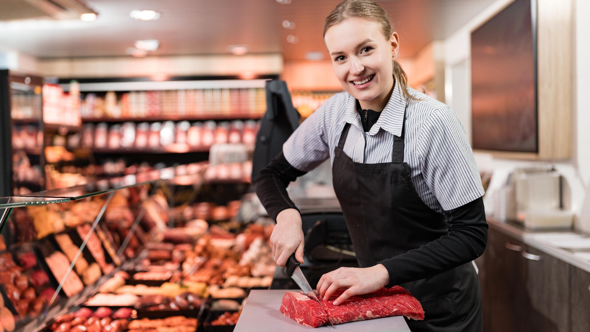 Ein lächelnder Metzger in Schürze schneidet ein Stück rohes Fleisch auf einem Schneidebrett an einer Fleischtheke, hinter deren Glas verschiedene Fleischsorten ausgestellt sind.