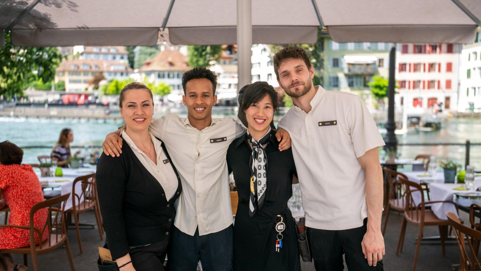 Vier Restaurantangestellte stehen lächelnd mit umschlungenen Armen im Freien unter einem Sonnenschirm, mit gedeckten Tischen und einer Stadtlandschaft am Fluss im Hintergrund.