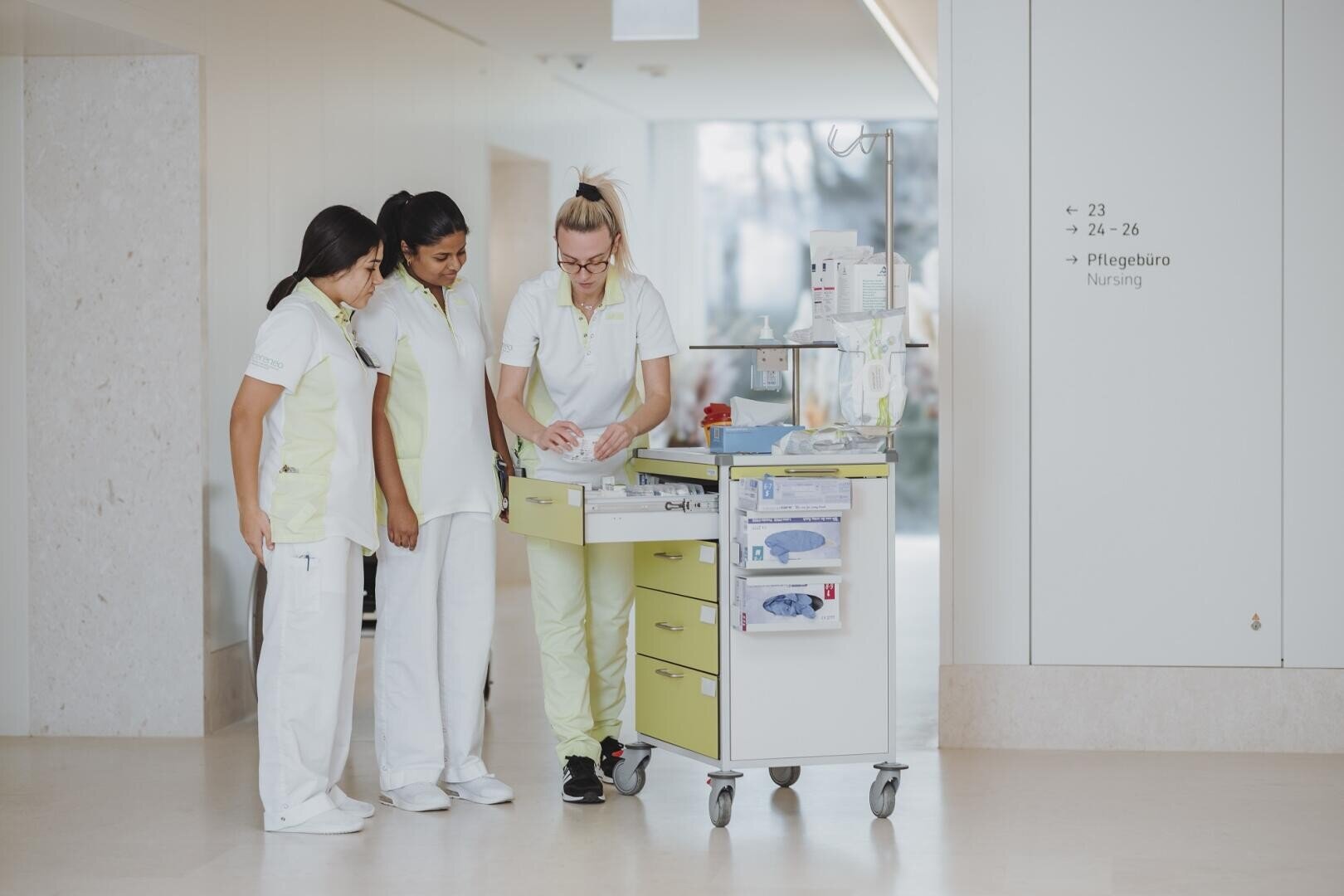 Drei Krankenschwestern in hellen Uniformen stehen neben einem Krankenwagen in einem Krankenhausflur. Eine Krankenschwester bereitet das Material vor, während die anderen zusehen. Ein Schild an der Wand weist den Weg zum Büro der Krankenschwester.