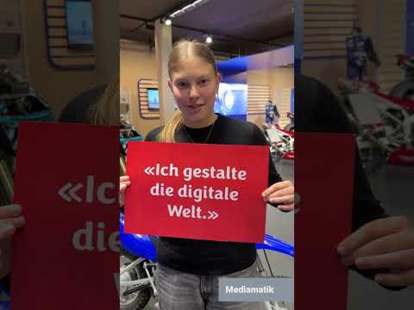Eine junge Person mit langen blonden Haaren hält ein rotes Schild mit der Aufschrift Ich gestalte die digitale Welt. Sie befinden sich in einem Innenraum, im Hintergrund sind Motorräder und Displays zu sehen.