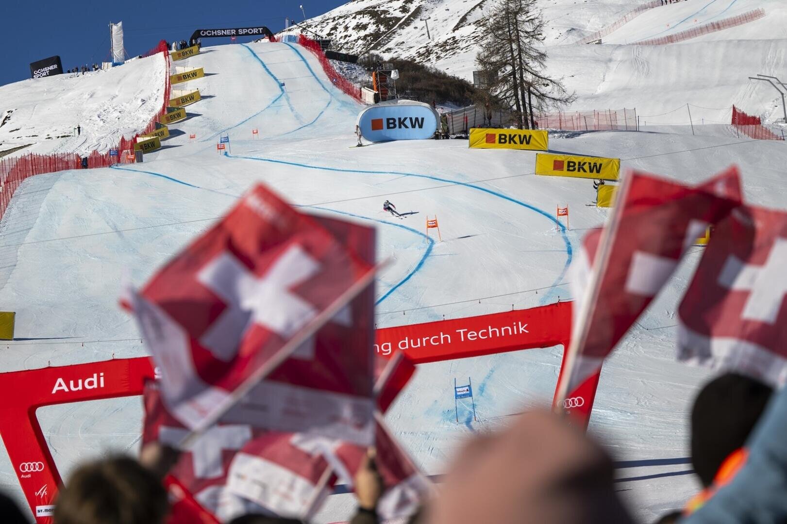 Zuschauer schwenken Schweizer Flaggen beim FIS Skirennen St. Moritz, während ein Skifahrer unter strahlend blauem Himmel eine verschneite Bergstrecke hinunterrast, die mit Toren und Bannern markiert ist.
