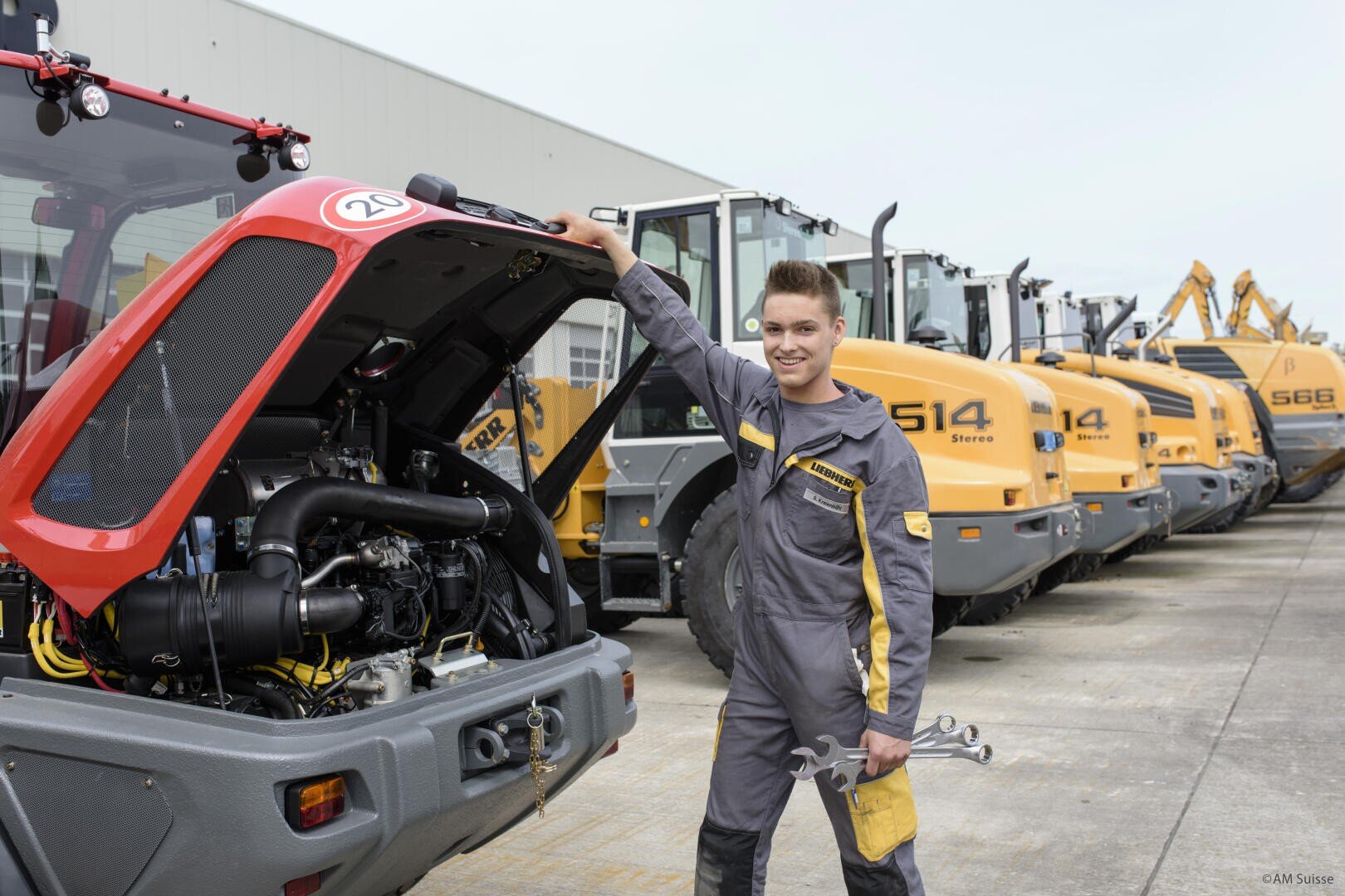 Ein lächelnder Mechaniker in einer grau-gelben Uniform steht neben einem roten Traktor mit offenem Motor und hält Werkzeuge in der Hand. Hinter ihm sind mehrere große gelbe Baufahrzeuge im Freien aufgereiht.