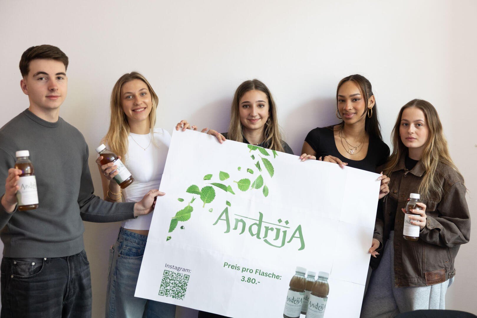 Fünf junge Erwachsene stehen in einem Raum und halten Flaschen und ein großes Plakat mit dem Markennamen Andrija und Produktdetails, einschließlich Preis und Links zu sozialen Medien. Die Atmosphäre wirkt locker und werblich.