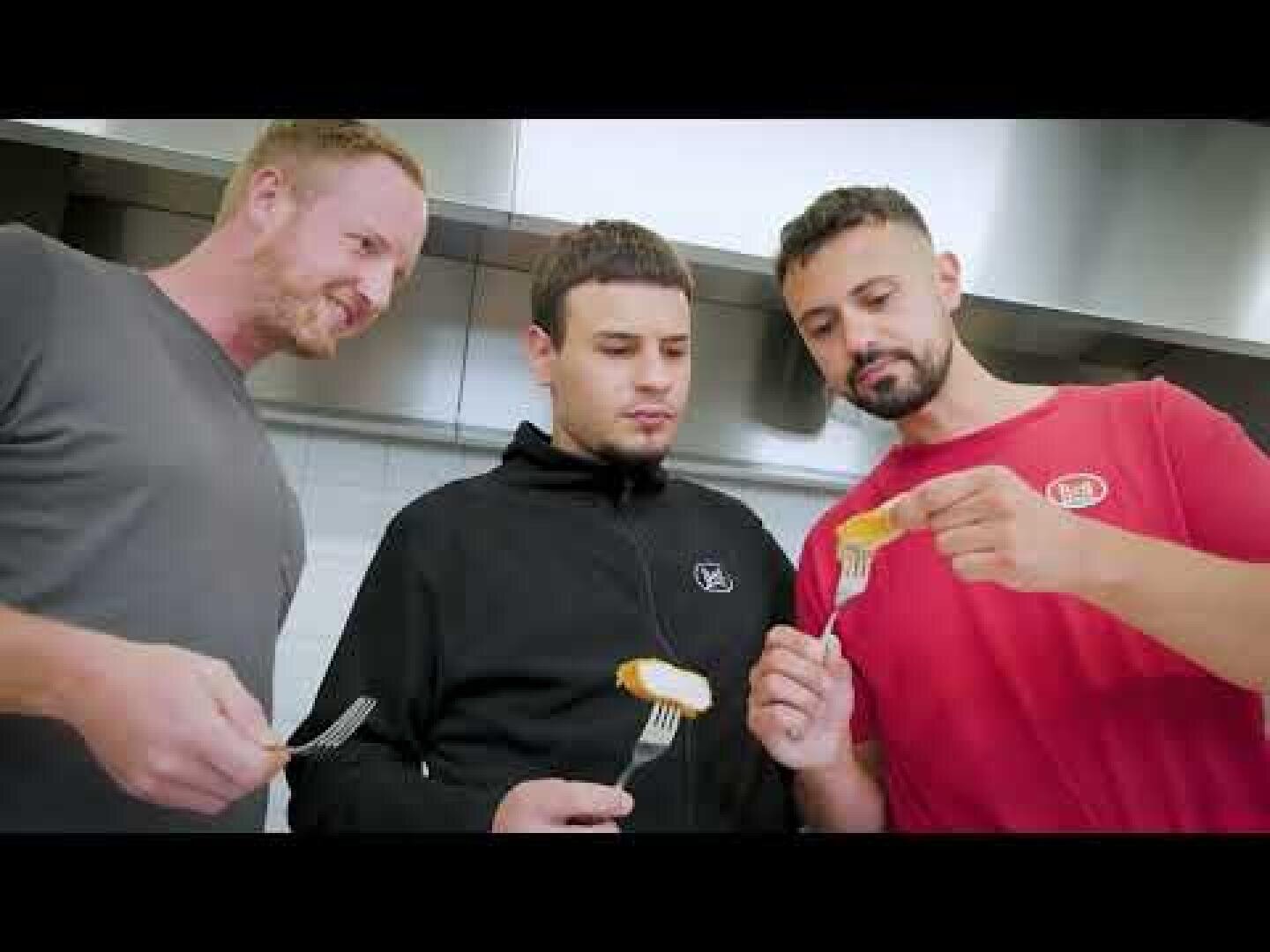 Drei Männer stehen in einer Küche und halten jeweils eine Gabel in der Hand. Der Mann auf der rechten Seite zeigt den beiden anderen, die ihn aufmerksam und interessiert beobachten, einen Bissen von seiner Gabel.