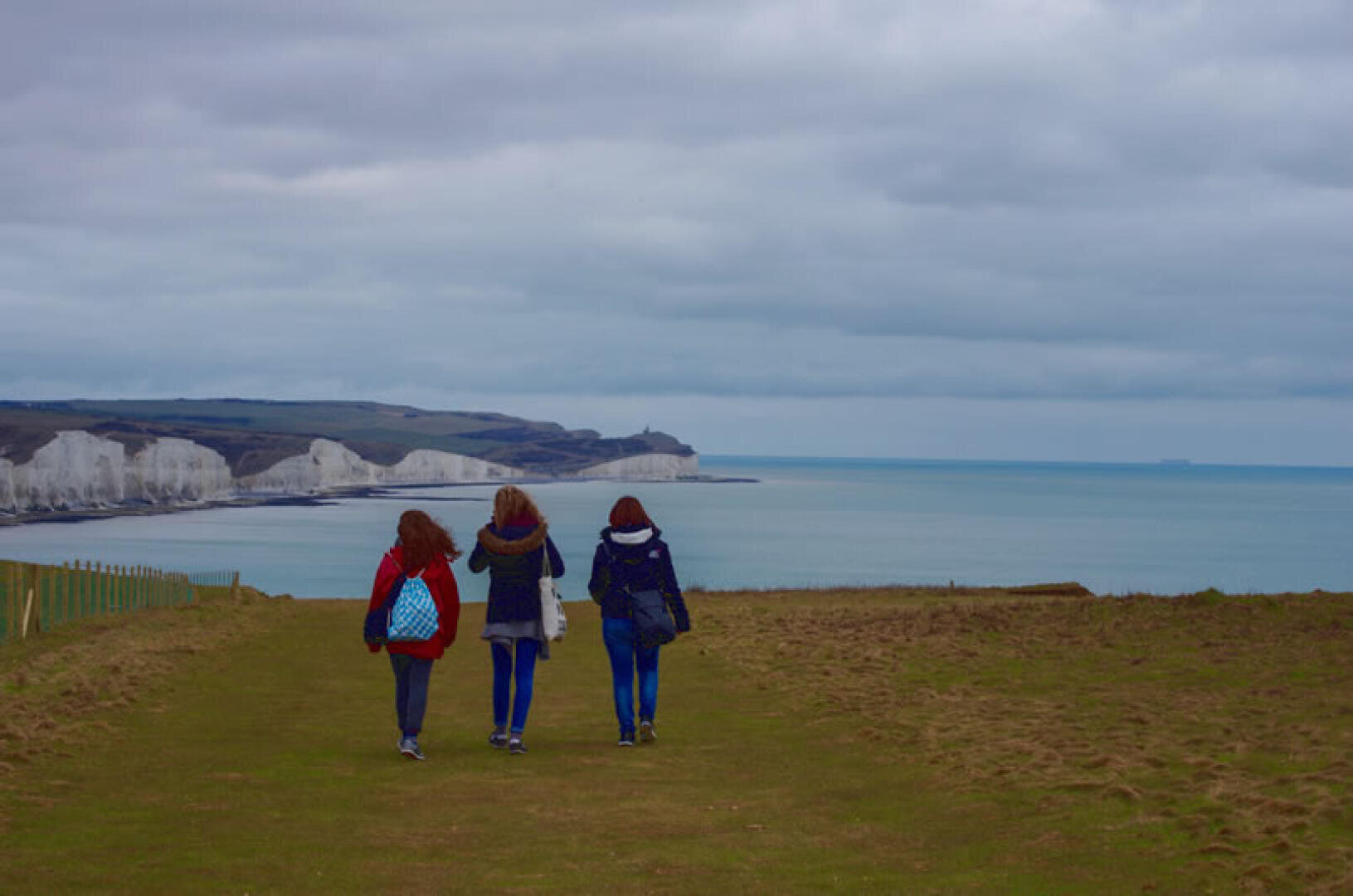 Drei Personen mit Rucksäcken gehen auf einem grasbewachsenen Weg in Richtung weißer Kreidefelsen am Meer unter einem bewölkten Himmel. Die Küste und die Klippen erstrecken sich in der Ferne auf der linken Seite.