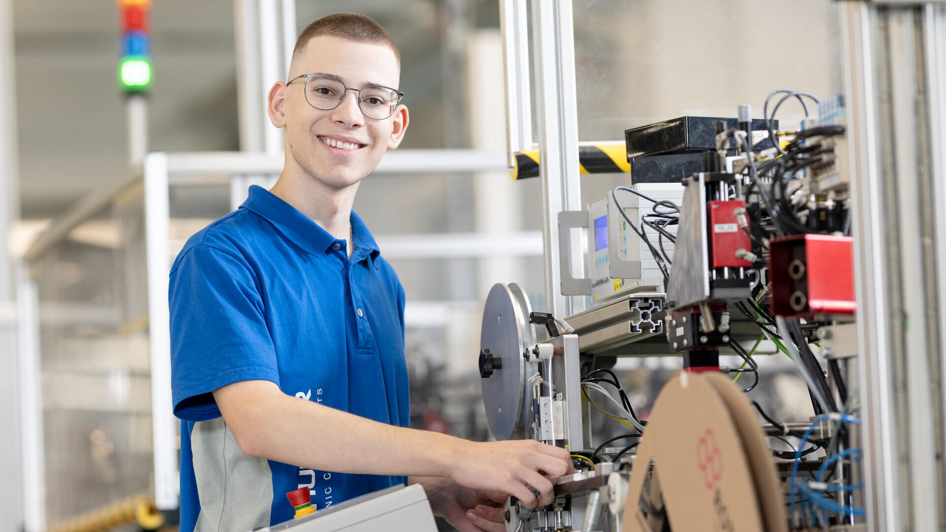 Ein junger Mann mit Brille und blauem Polohemd bedient Maschinen in einer modernen Industrieumgebung. Er lächelt in die Kamera, während er Geräte einstellt, die von Kabeln und elektronischen Bauteilen umgeben sind.