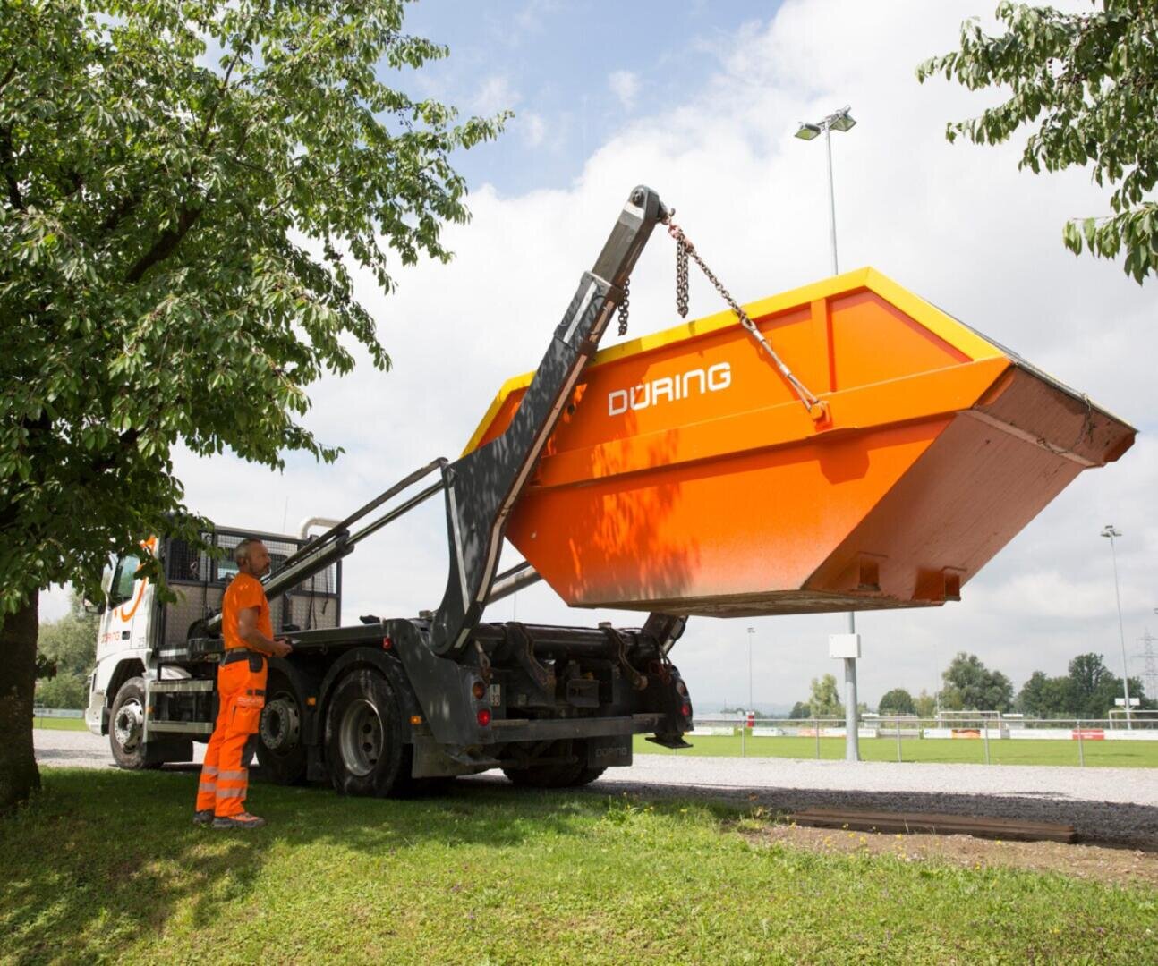 Ein Arbeiter in orangefarbener Sicherheitskleidung bedient einen Autokran, um einen orangefarbenen Müllcontainer mit der Aufschrift DüRING auf eine Wiese in der Nähe eines Baumes und einer Freifläche zu heben.