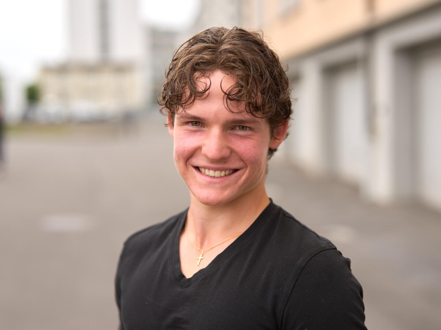 Ein junger Mann mit lockigem braunem Haar in einem schwarzen Hemd lächelt im Freien und trägt eine goldene Kreuzkette. Der Hintergrund ist unscharf und zeigt Gebäude und eine Straße.