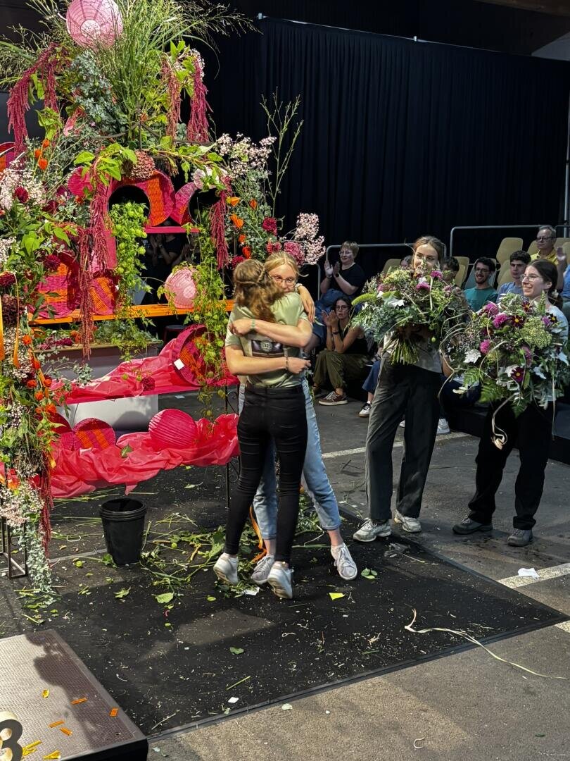 Zwei Menschen umarmen sich neben einem großen, farbenfrohen Blumenarrangement auf der Bühne, während zwei weitere, die Blumensträuße in den Händen halten, lächelnd zuschauen. Im Hintergrund sitzen Zuschauer.