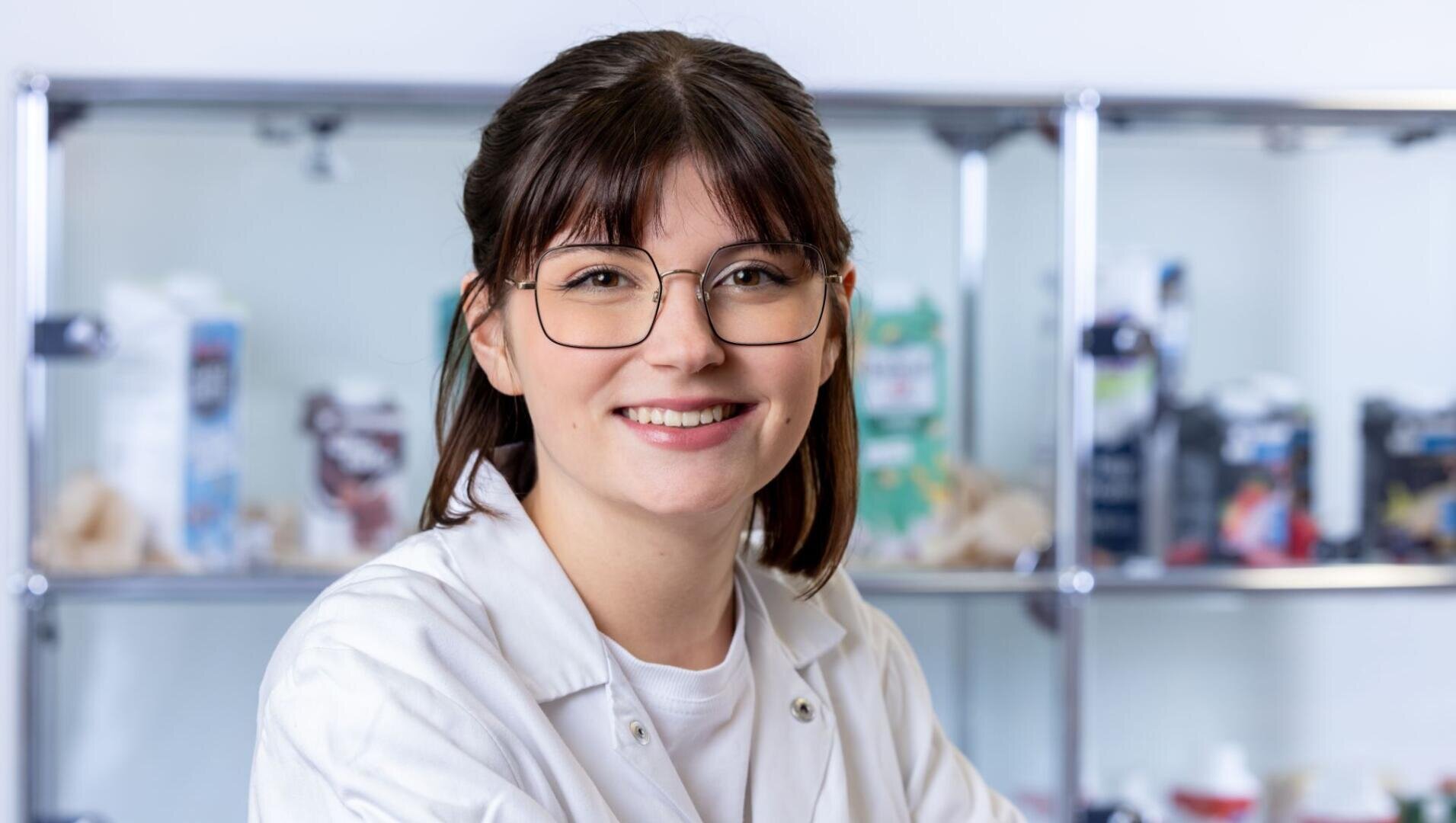 Eine junge Frau mit Brille und weißem Laborkittel steht lächelnd in einem Labor, im Hintergrund sind Regale mit verschiedenen Behältern und Verpackungen zu sehen.