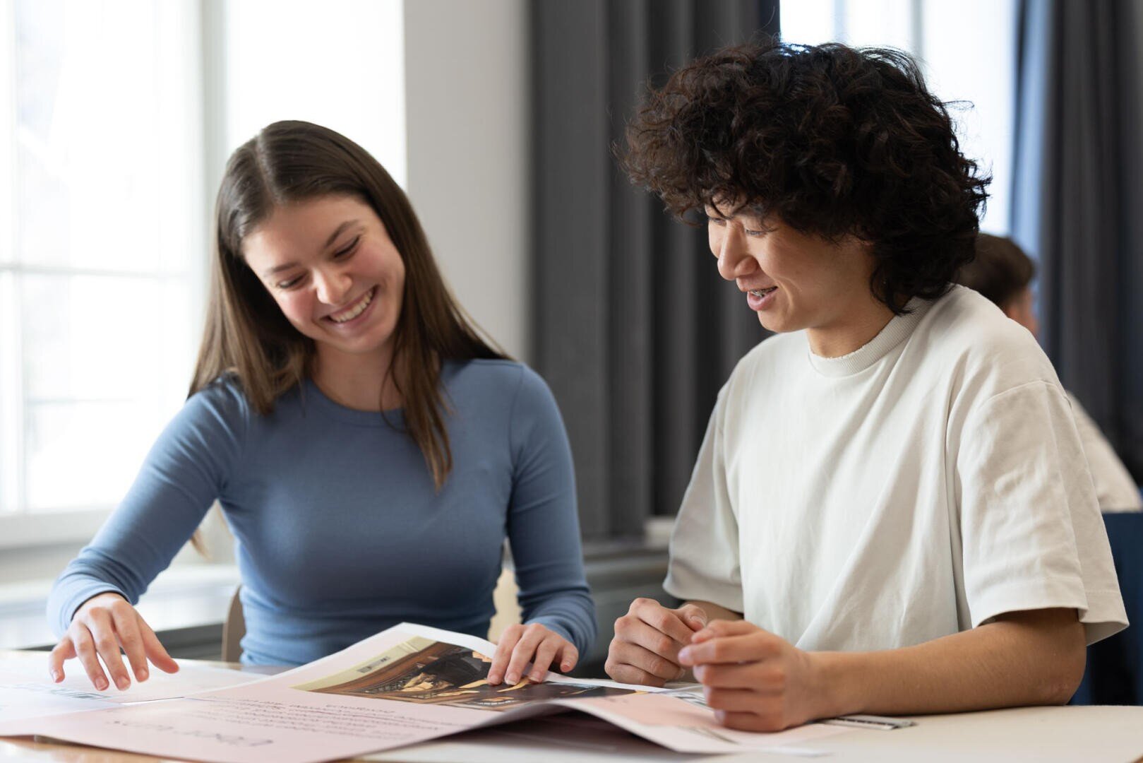 Zwei junge Erwachsene sitzen lächelnd an einem Tisch und betrachten gemeinsam große Papiere oder Plakate in einem hellen Raum, was auf eine Arbeits- oder Lernumgebung schließen lässt.