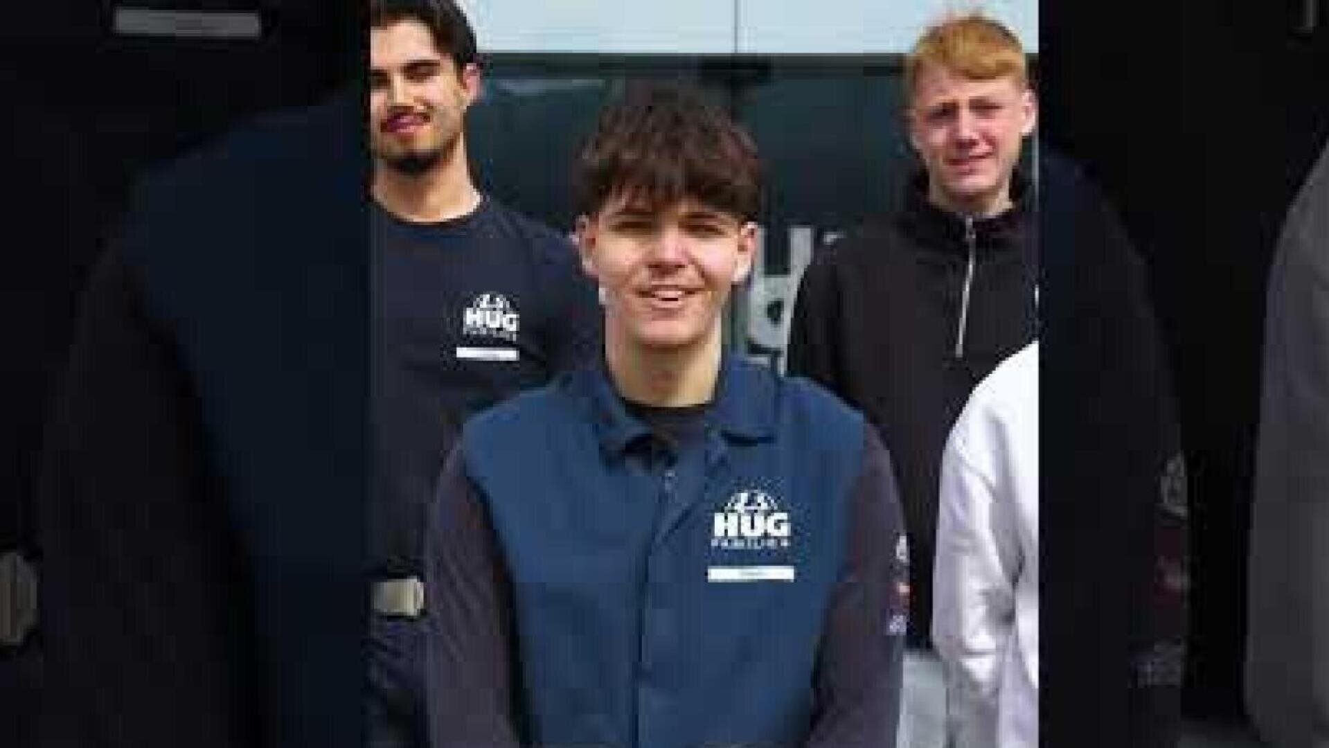 Drei junge Menschen stehen beieinander und lächeln in die Kamera. Die Person ganz vorne trägt eine dunkelblaue Weste mit der Aufschrift „HUG“, während die beiden dahinter dunkle Hemden tragen. Sie scheinen sich im Freien zu befinden, möglicherweise vor einem Gebäude.