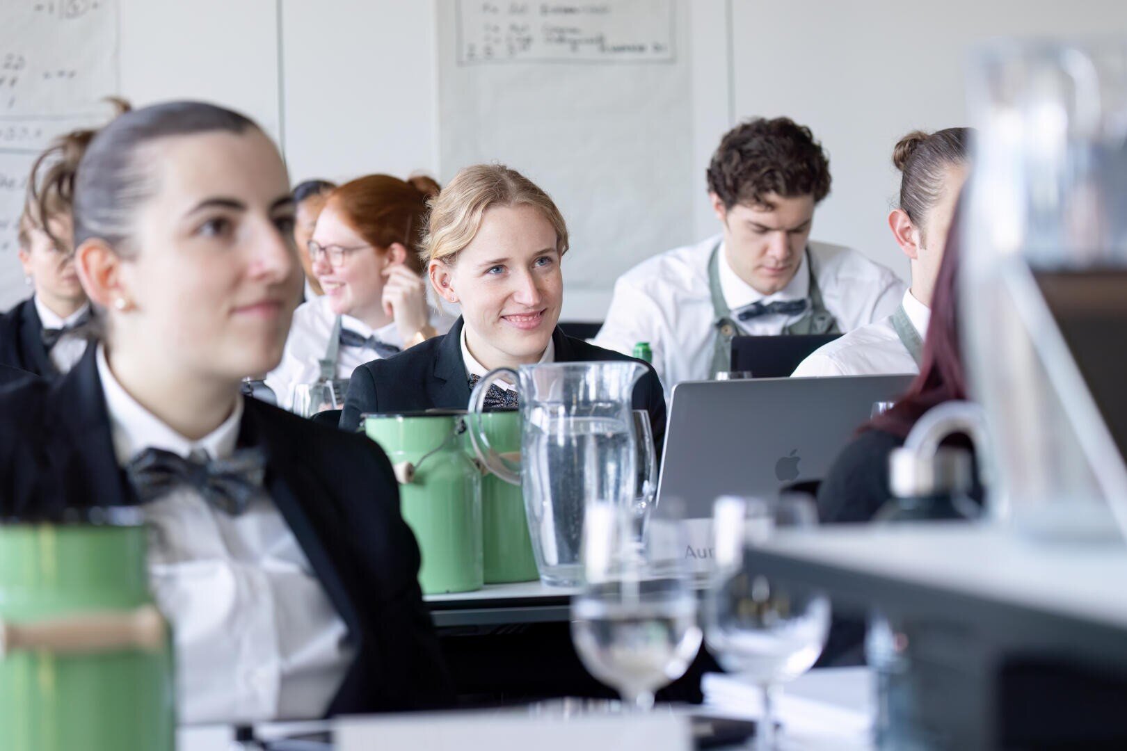 Eine Gruppe junger Erwachsener in formeller Kleidung mit Fliege sitzt an den Tischen in einem Klassenzimmer und hört aufmerksam zu. Eine Frau in der Mitte lächelt, und auf den Tischen sind Krüge und Gläser mit Wasser zu sehen.