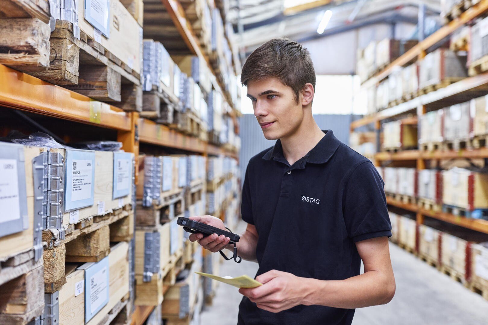 Ein junger Mann in einem schwarzen Poloshirt hält einen Barcode-Scanner und einen Notizblock in der Hand, während er in einem Lagerhaus arbeitet, umgeben von Regalen voller Kisten und Paletten.