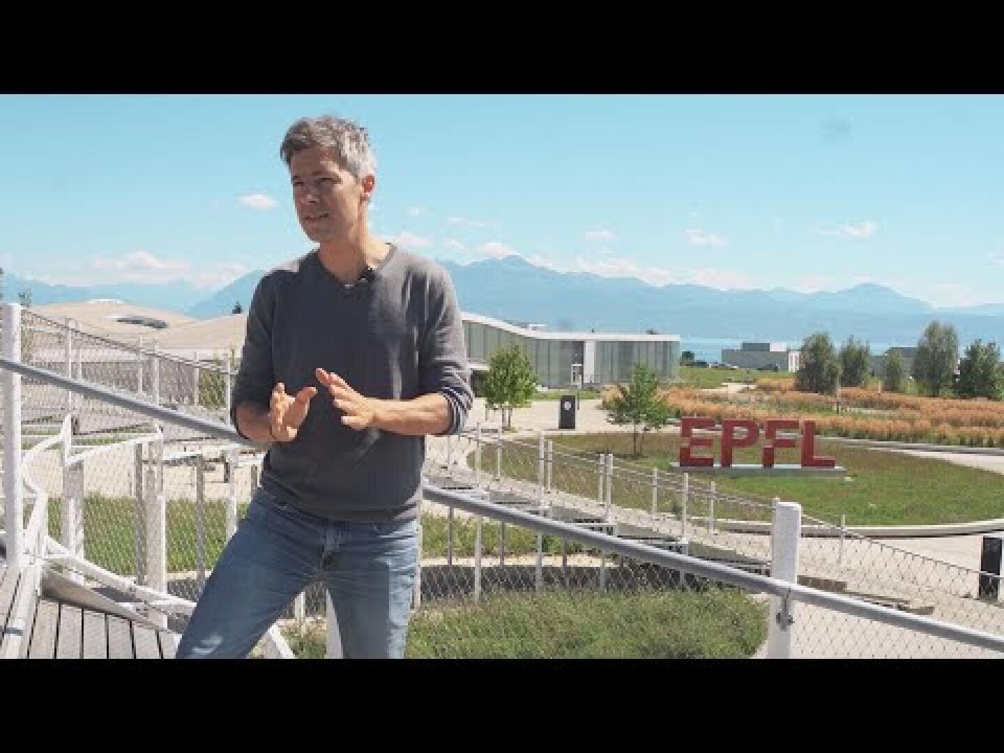 Ein Mann in einem grauen Pullover gestikuliert im Freien auf einer Rampe in der Nähe einer Wiese. Im Hintergrund sind das rote EPFL-Schild und die Berge unter einem klaren blauen Himmel zu sehen.