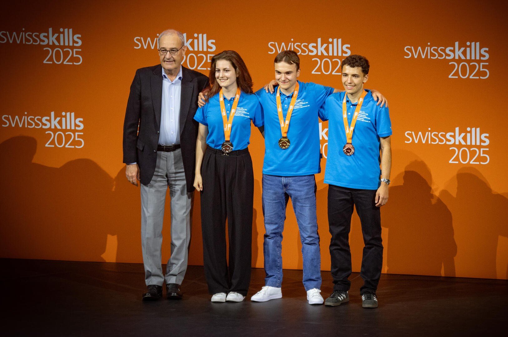 Vier Personen stehen vor einer orangefarbenen Kulisse der SwissSkills 2025. Drei junge Menschen in blauen Hemden tragen Medaillen und legen lÀchelnd die Arme umeinander, wÀhrend ein Àlterer Mann im Anzug daneben steht.