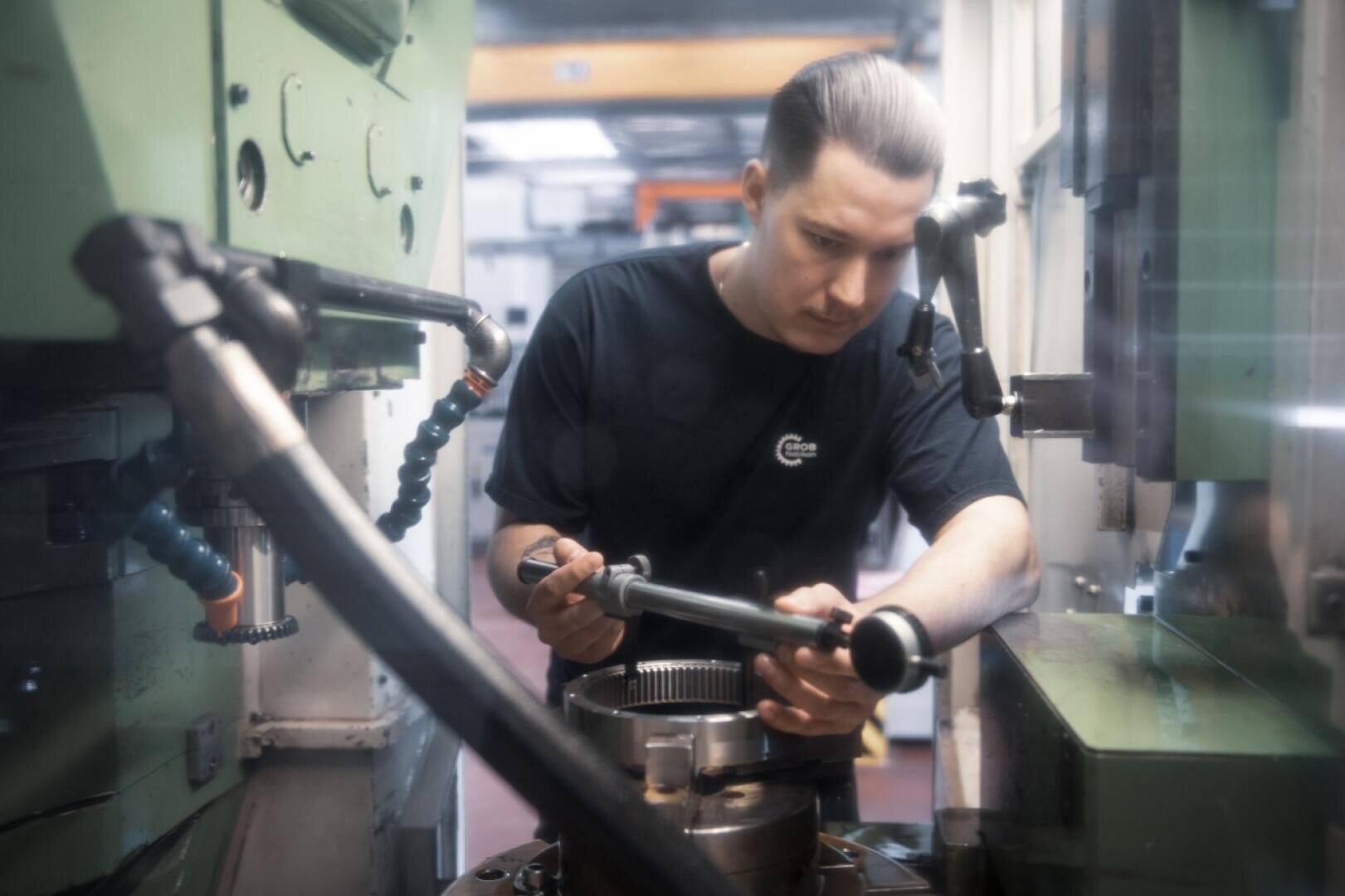 Eine Person in einem dunklen Hemd bedient eine Maschine und justiert ein Metallteil in einer industriellen Werkstattumgebung mit grüner Ausrüstung und sichtbaren mechanischen Bauteilen.