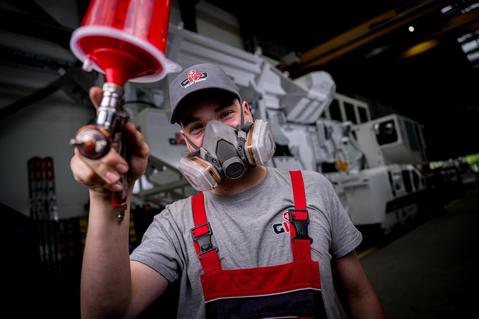 Ein Arbeiter, der eine Atemschutzmaske, einen rot-grauen Overall und eine Kappe trägt, hält eine Farbspritzpistole, die auf die Kamera in einer Industrieanlage mit großen Maschinen im Hintergrund gerichtet ist.