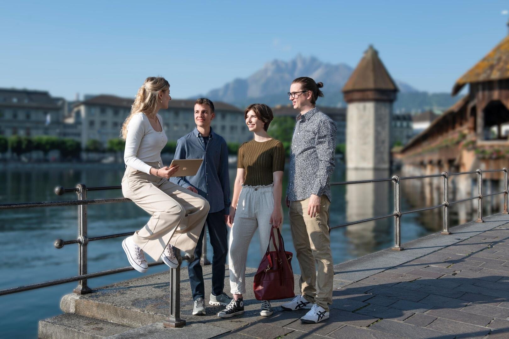 Vier junge Erwachsene unterhalten sich und lächeln an einem Geländer am Seeufer in einer europäischen Stadt, mit historischen Gebäuden, einer Holzbrücke und Bergen im Hintergrund an einem sonnigen Tag. Eine Person hält ein Tablet, während sie auf dem Geländer sitzt.