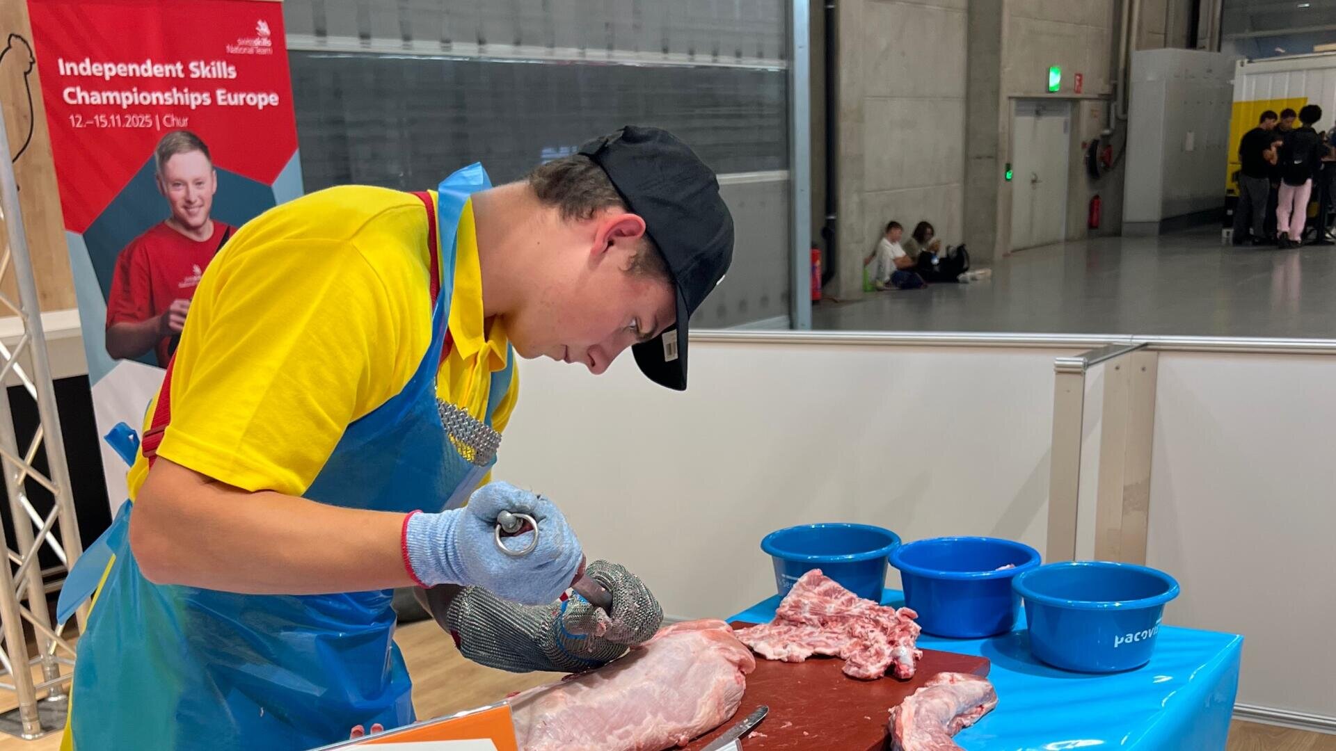 Ein junger Mann in Schutzkleidung bereitet an einem Wettbewerbsstand Fleisch zu; neben ihm stehen blaue Schüsseln. Im Hintergrund sind ein Banner der „Skills Championships Europe“ und sitzende Personen zu sehen.