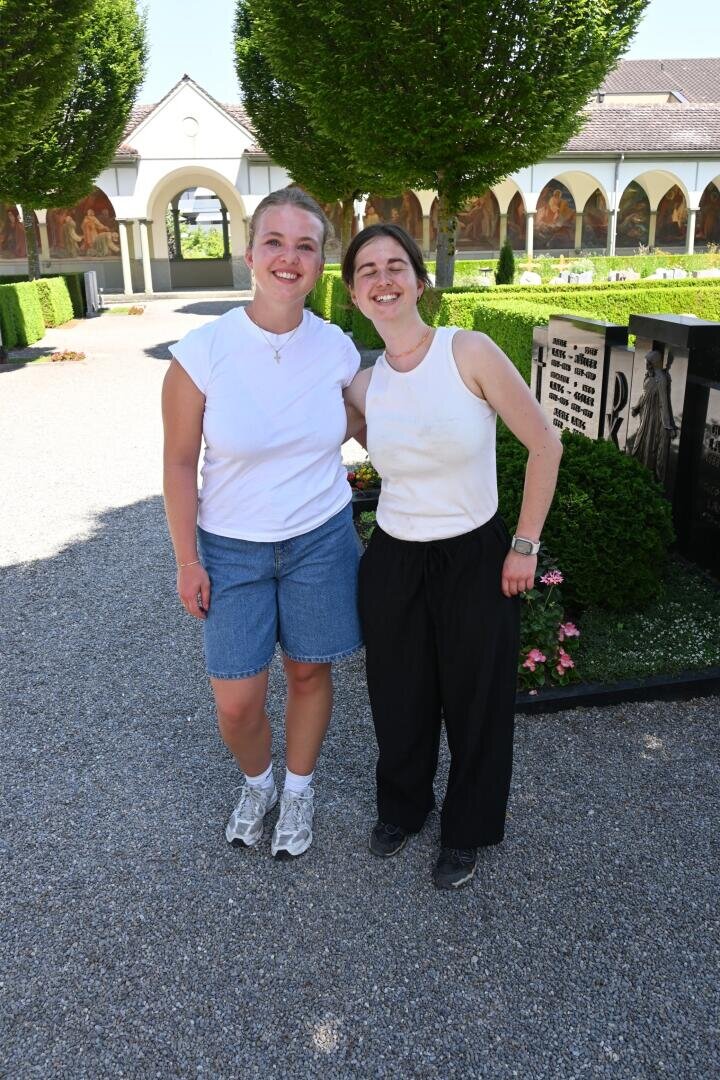 Zwei Frauen stehen lächelnd nebeneinander im Freien auf einem Schotterweg, im Hintergrund sind grüne Hecken, Bäume und ein Gebäude mit Bögen und Wandmalereien zu sehen. Die eine trägt Shorts und Turnschuhe, die andere eine schwarze Hose und dunkle Schuhe.