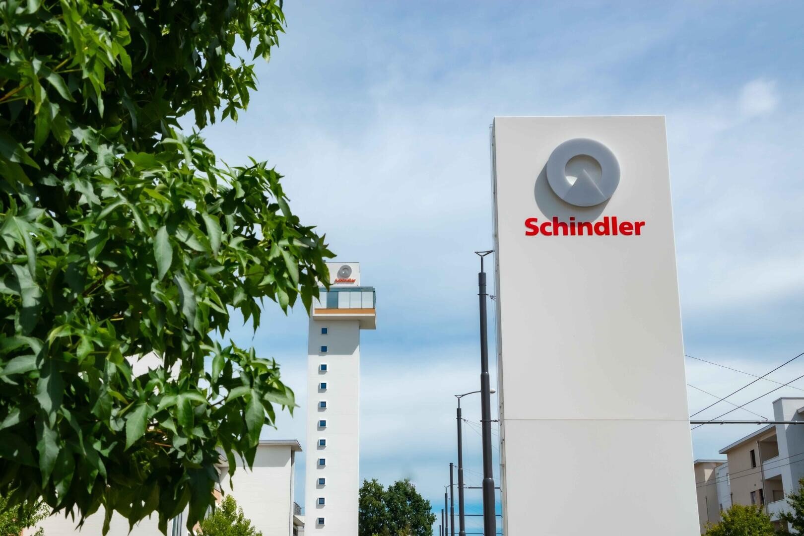 Vor einem teilweise bewölkten Himmel steht draußen in der Nähe von Bäumen und einem modernen Gebäude ein großes weißes Schindler-Schild mit einem roten Logo.