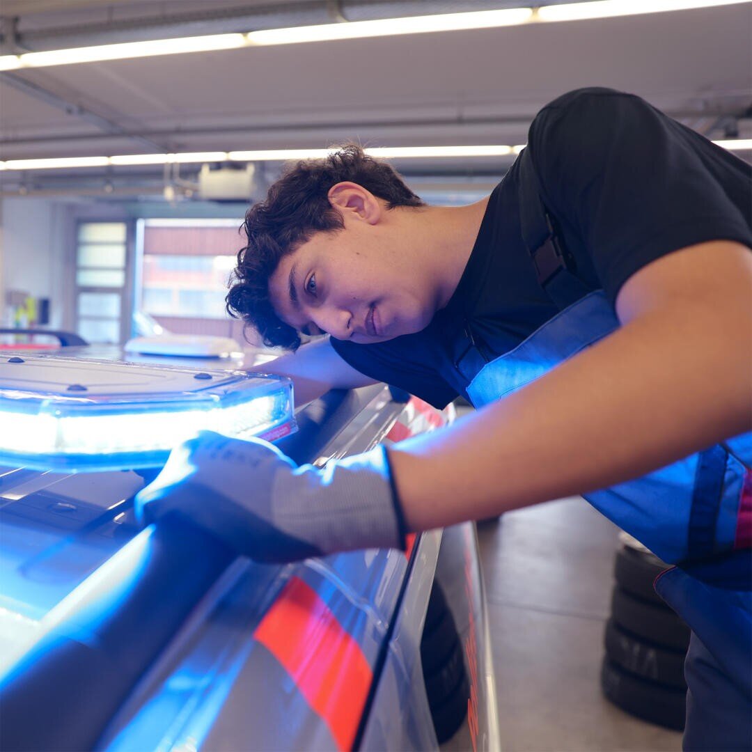 Ein junger Mechaniker mit Handschuhen prüft das blinkende Blaulicht auf dem Dach eines Fahrzeugs in einer hell erleuchteten Werkstatt oder Garage.