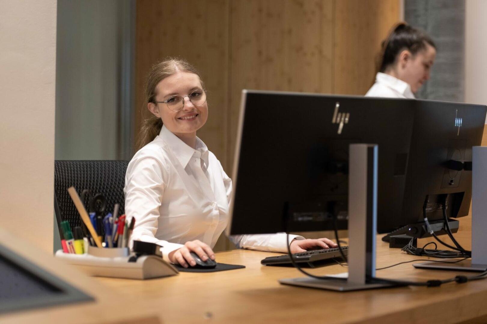 Eine Frau mit weißem Hemd und Brille lächelt an einem Schreibtisch mit Computer und Büromaterial, während im Hintergrund eine andere Person an einem Computer arbeitet.