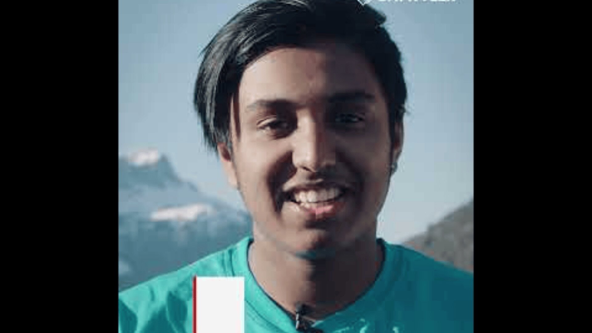 Eine junge Person mit dunklen Haaren lächelt in die Kamera. Sie steht im Freien mit Bergen und einem blauen Himmel im Hintergrund. Die Person trägt ein blaugrünes Hemd.