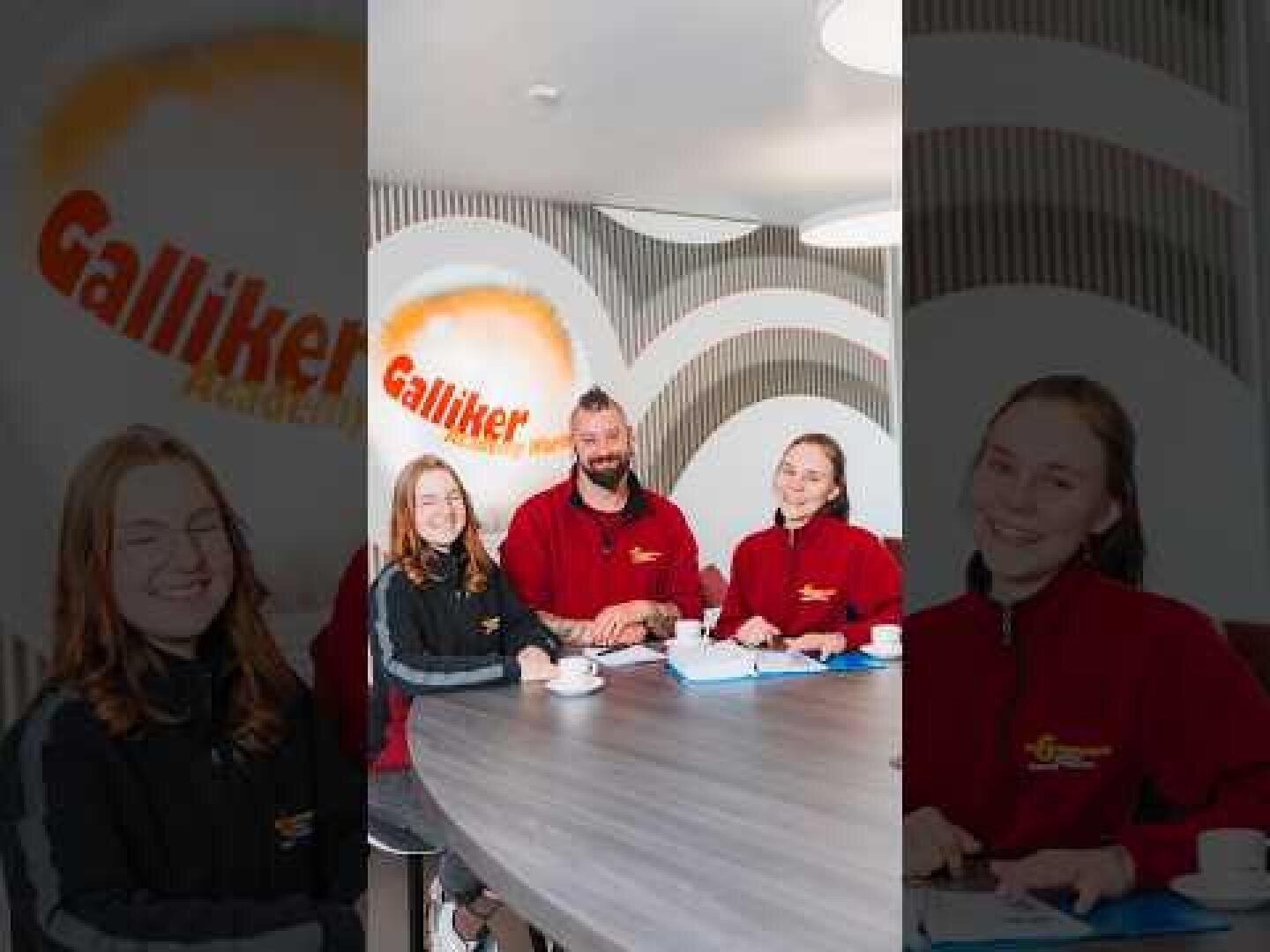 Vier Personen in roten und schwarzen Uniformen sitzen lächelnd mit Unterlagen und Kaffeetassen um einen runden Tisch herum. Im Hintergrund ist eine Wand mit dem orangefarbenen Galliker-Logo und geschwungenen Designelementen zu sehen.