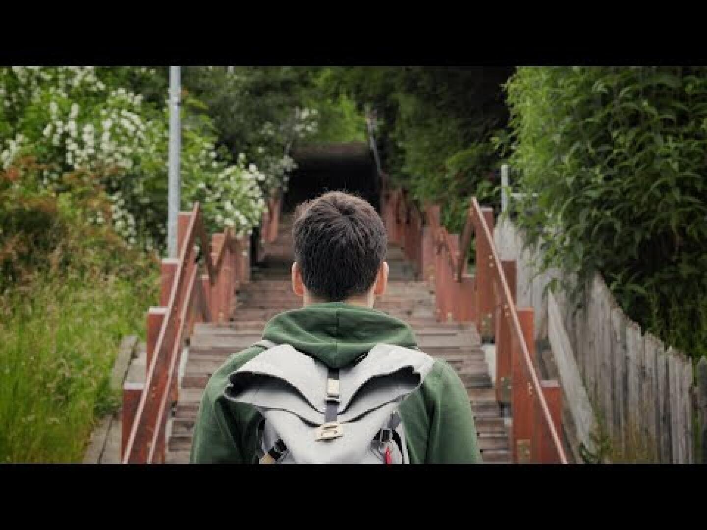 Eine Person mit kurzen, dunklen Haaren und einem grauen Rucksack geht eine hölzerne Außentreppe hinauf, die von Grünpflanzen und Büschen umgeben ist.